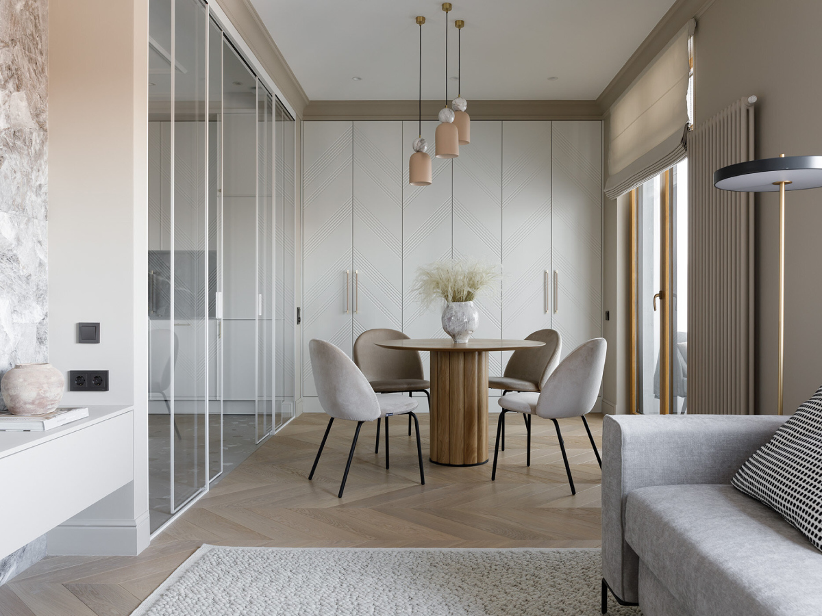 Soft neutrals and layered textures create a serene dining nook. A round wood table adds warmth, while curved chairs invite lingering. Pale pendants with marble accents bring a touch of whimsy, and subtly patterned cabinet doors add texture to the space.