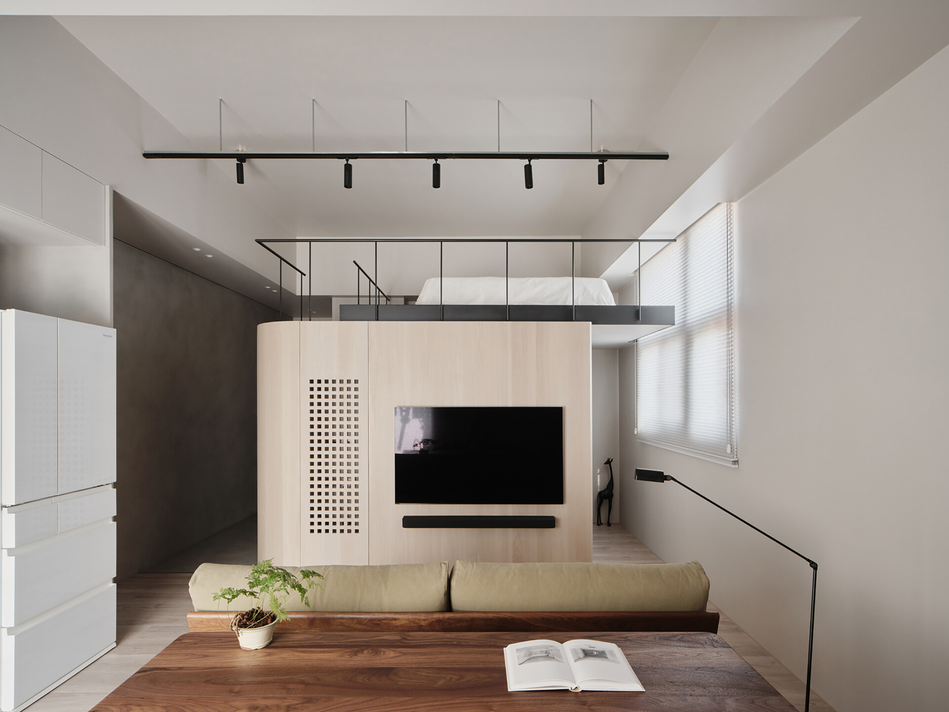 Soft, pale wood tones and clean lines define this clever loft setup, where every inch is thoughtfully designed. The built-in structure combines a TV wall, concealed storage, and a raised sleeping area above, all bathed in gentle natural light. Matte black railings and track lighting add a sharp contrast, giving the space a quietly modern edge. It’s a smart solution that feels both minimal and cozy, perfect for small-space living without ever feeling cramped.